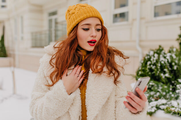 雪街上站着一个漂亮的红发女人 手里拿着手机穿着外套 戴着帽子 手里拿着智能手机的可爱的姜黄色女士深色冻结冬季