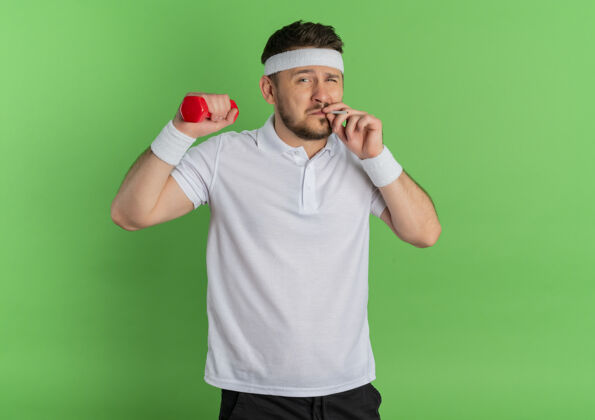 健身身穿白衬衫 头箍 举着哑铃 做运动 抽着烟的年轻健身男子 不良习惯的体育理念屹立在绿色的背景之上男士习惯锻炼