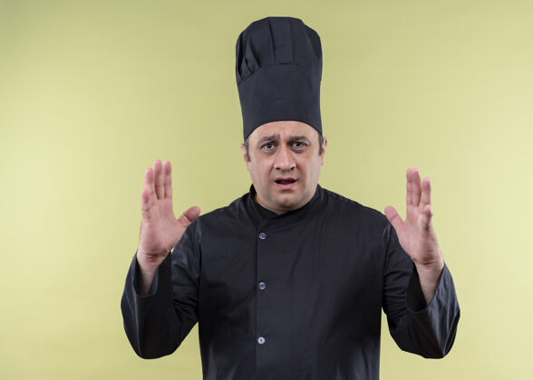 厨师男厨师长身穿黑色制服 头戴厨师帽 双手示意大尺寸标志 惊讶地站在绿色背景上测量标志制服男性穿着