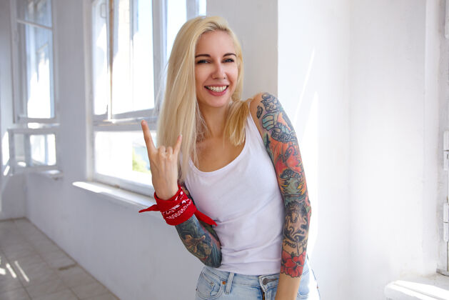 情感室内拍摄的年轻金发长发女子纹身做摇滚标志与提高手和微笑愉快地在前面 摆在大窗口前微笑摇滚乐穿