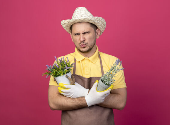 男人年轻的园丁穿着连体衣 戴着帽子 戴着工作手套 手里拿着盆栽植物 眉头紧锁 严肃地站在粉红色的墙上 望着前方男皱眉粉色