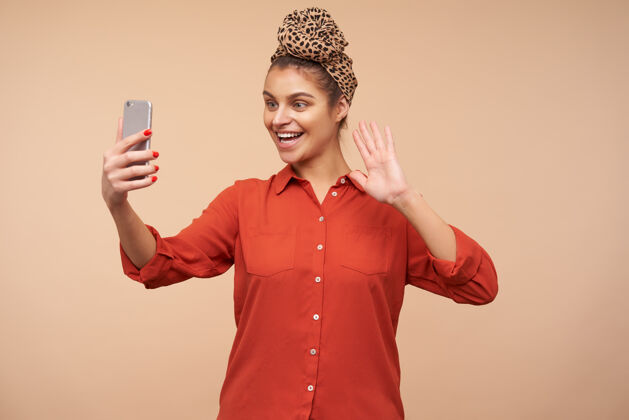 头巾快乐的年轻可爱的棕色头发的女人 戴着头巾 在手机前愉快地微笑 举起手打招呼 一边打视频电话 在米色的墙上摆姿势化妆品女性美女