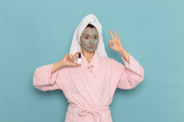 肖像正面图身着粉色浴袍的年轻女性淋浴后拿着指甲油在蓝色墙壁上美丽的清水自理淋浴抛光淋浴尺子