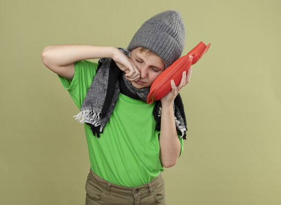 不适生病的小男孩穿着绿色t恤 戴着暖和的围巾和帽子 感觉不舒服 拿着水瓶取暖 忍受着站在光墙上流鼻涕的痛苦流涕温暖持有