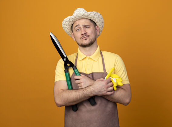 人年轻的园丁穿着连体衣 戴着帽子 戴着工作手套和树篱剪 站在橙色的墙上自信地看着前方手套篱笆人