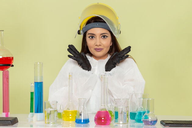 成人正面图身着特殊防护服的年轻女化学家坐在桌子前 拿着绿色墙壁上的溶液工作化学实验室化学女科学科学年轻人