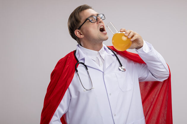 男人超级英雄医生身穿白大褂 披着红斗篷 戴着听诊器的眼镜 站在白墙上拿着装有黄色液体的烧瓶喝酒听诊器超级英雄外套