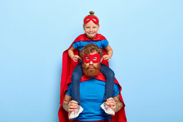 家庭滑稽的小女孩穿着超级英雄的服装 坐在爸爸的肩膀上 让他的耳朵突出 和爸爸玩得很开心为人父母触摸玩耍