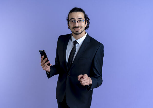 男人身穿黑色西装 戴着眼镜的商务人士展示着智能手机 手指指着前面 微笑着自信地站在蓝色的墙上商人站立人