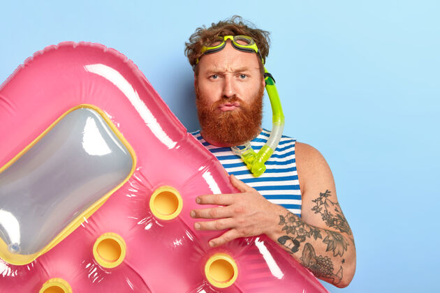阳刚不高兴的胡子家伙摆着粉红色的气垫 戴着泳镜和潜水面具游泳池携带潜水