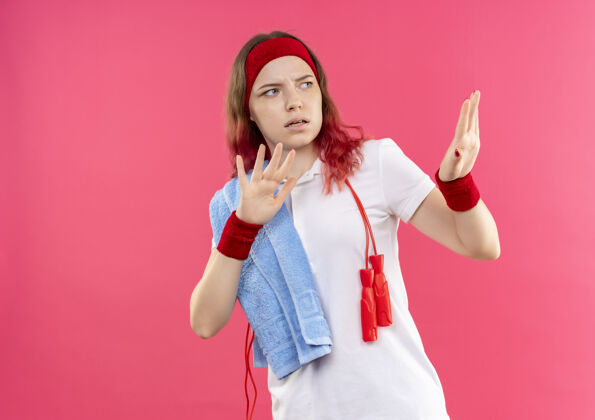 防御戴着头巾 肩上扛着毛巾的年轻运动型女子站在粉红色的墙上做着防御手势 双手带着恐惧的表情看着人恐惧人