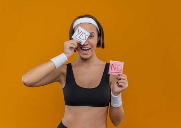 表演身穿运动服 头箍的年轻健身女士展示着两张写着“是”和“否”的提醒纸 她微笑着用纸遮住一只眼睛站在橙色的墙上头带眼睛运动