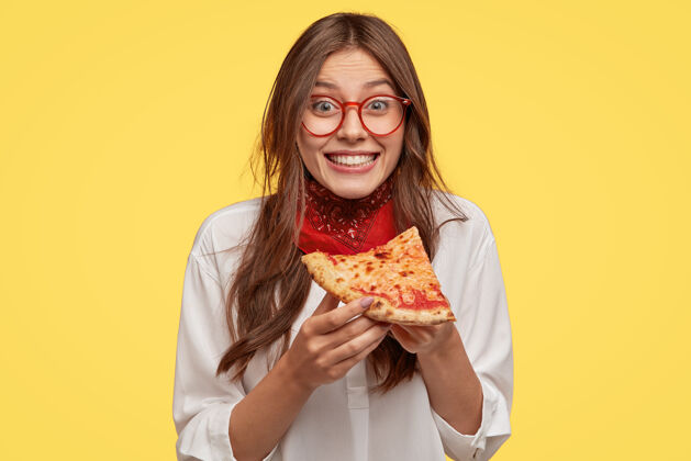 成人满意的女人拿着一块披萨的照片 和朋友在披萨店闲暇时感到高兴 开心地直接穿上休闲装 隔着黄墙午餐头巾白种人块