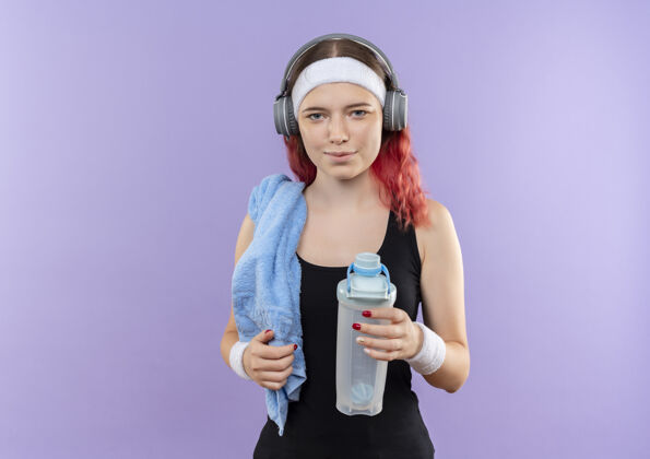 年轻人年轻的健身女孩头戴耳机 脖子上戴着毛巾 手里拿着一瓶水 微笑着站在紫色的墙上瓶子头水