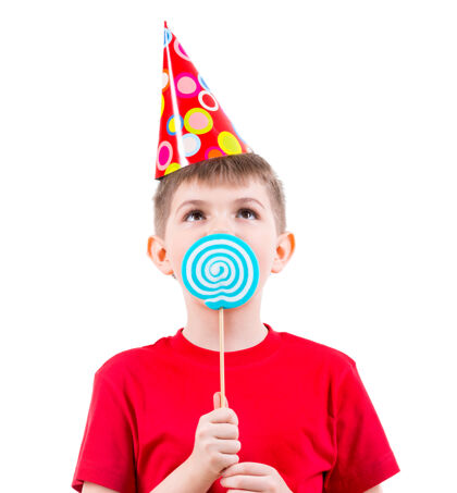 帽子小男孩穿着红色t恤 戴着派对帽 吃着彩色糖果-白色的隔离带积极高兴漂亮