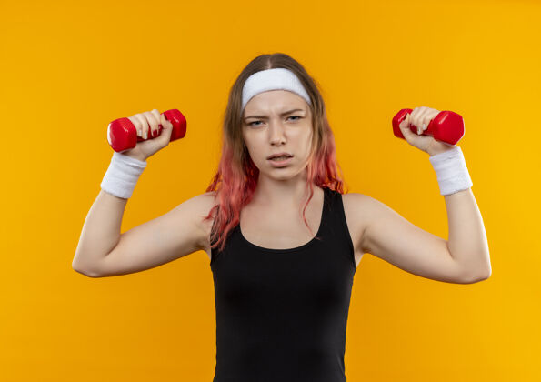 运动装穿着运动服的年轻健身女士正脸站在橙色的墙上 用哑铃做运动哑铃看健康