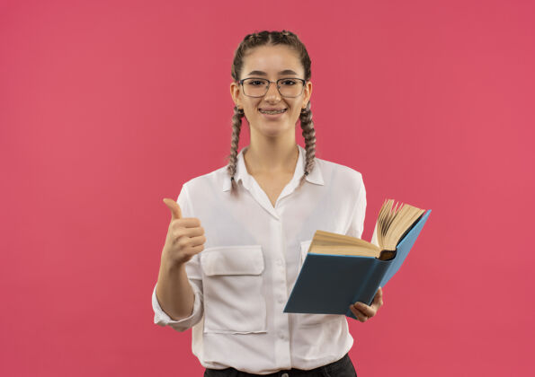 学生戴着眼镜 梳着辫子 穿着白衬衫的年轻女学生 拿着打开的书 微笑着站在粉色的墙上 竖起大拇指望着前面开放人显示