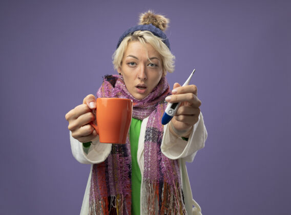 帽子有病的不健康的女人 短头发 戴着暖和的围巾和帽子 感觉不舒服 拿着一杯热茶和温度计 站在紫色背景上看着摄像机体温计不健康热