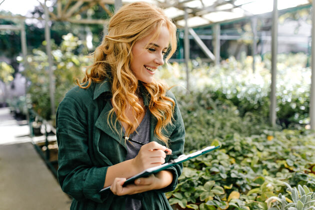 成人一位金发碧眼 笑容温柔的年轻女子 身着绿色长袍 系着腰带 正在温室里工作公园女性花园