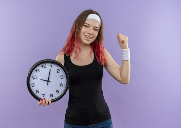女人身穿运动服的年轻健身女士手持挂钟 握紧拳头 高兴地站在紫色的墙上走了出来人握紧运动员