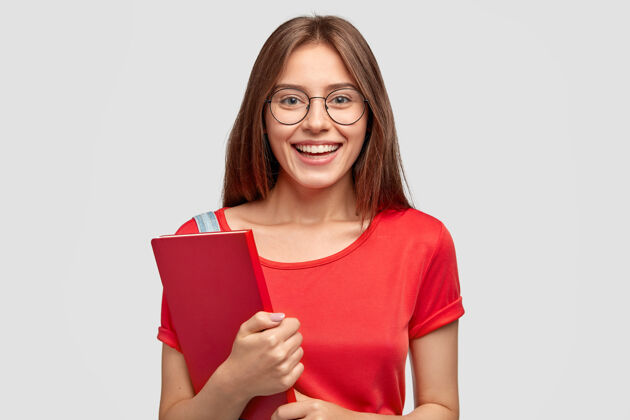 室内积极的白人女孩 微笑迷人 穿着红色t恤 拿着课本 靠着白墙做模特 有学习的心情 戴着眼镜 有良好的视力青春 学习的理念情感青少年白种人