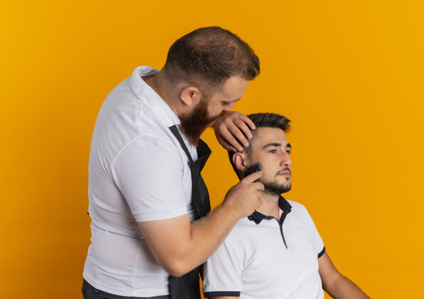 刮胡子专业的胡须理发师围着围裙用剃须刀机给站在橘色墙上的年轻人理发围裙做胡子机器