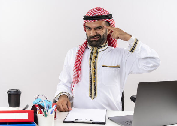 眨眼身着传统服装的阿拉伯商人坐在桌旁 用笔记本电脑眨眼 指着他的太阳穴专注于办公室里的一项工作任务寺庙桌子