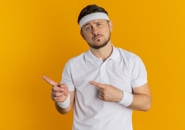 悲伤身穿白衬衫 头戴头巾的年轻健身男子面带悲伤的表情 手指指向站在橙色墙上的一侧男人头带表情