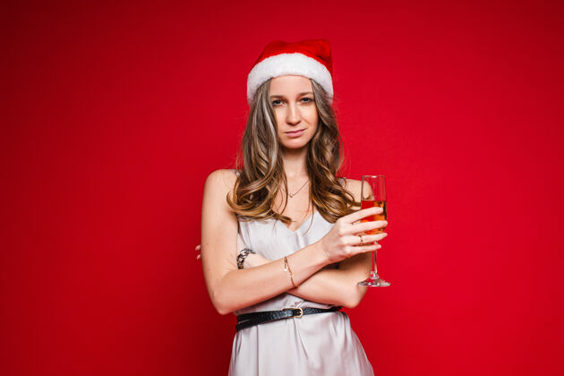 女士戴着圣诞帽 穿着节日礼服 手持香槟酒杯在红色背景上摆姿势的年轻漂亮女性 复制空间美丽圣诞老人女人