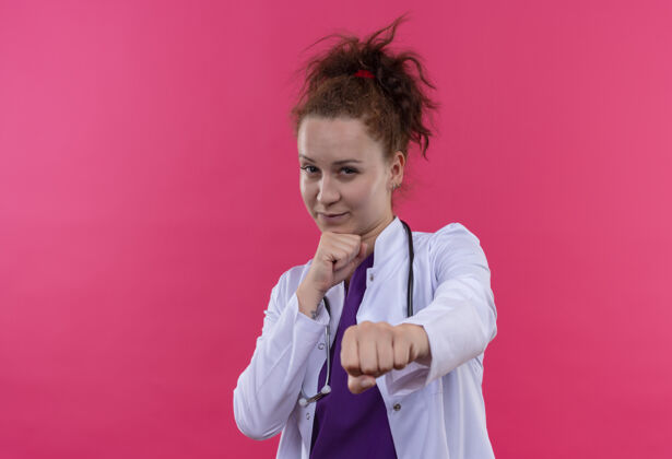 听诊器年轻的女医生穿着白大褂 手持听诊器 用拳头指着粉色的墙上 看上去很自信女人看医生