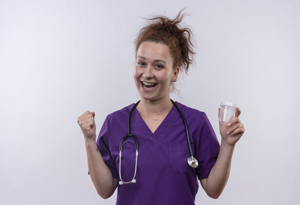 医生年轻的女医生穿着医疗制服 手持听诊器 高兴地伸出握紧的拳头站在白墙上女人罐子拳头