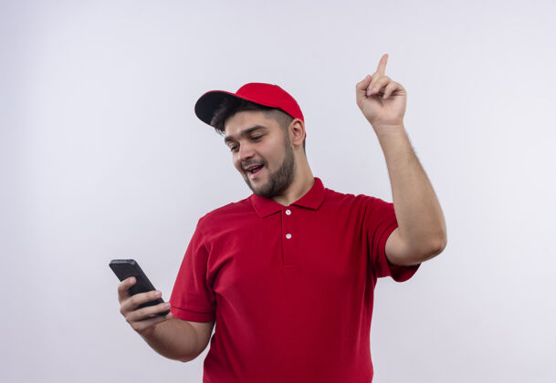 送货年轻的送货员穿着红色制服 戴着帽子 看着他的智能手机屏幕 用手指着 有个好主意屏幕男人向上