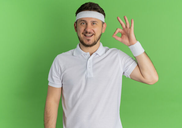 教练身穿白衬衫 头戴花环的年轻健身男子站在绿色的墙上 面带微笑 高高兴兴地唱着歌头带男站着