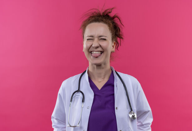 听诊器年轻的女医生身穿白色外套 手持听诊器 高兴地伸出舌头站在粉红色的墙上穿女人外套