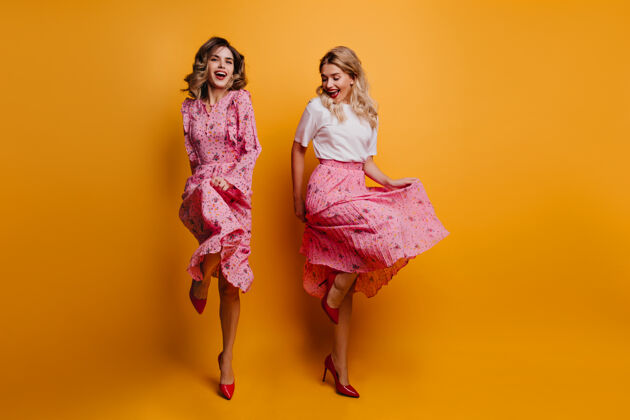 朋友苗条兴奋的女孩和朋友跳舞 温文尔雅的女士们穿着粉色的衣服在黄色的墙上一起冷娱乐时尚享受