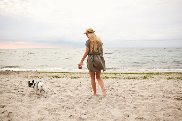 情绪户外照片 苗条的年轻女性 金色长发 穿着夏装 头戴水手帽 在灰蒙蒙的阴天 牵着狗在沙滩上散步白色阴天姿势