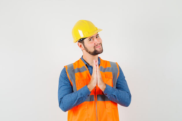 头盔年轻的建设者穿着衬衫 背心 头盔 手拉手 做着祈祷的手势 看上去很乐观 正面看去祈祷手勤杂工