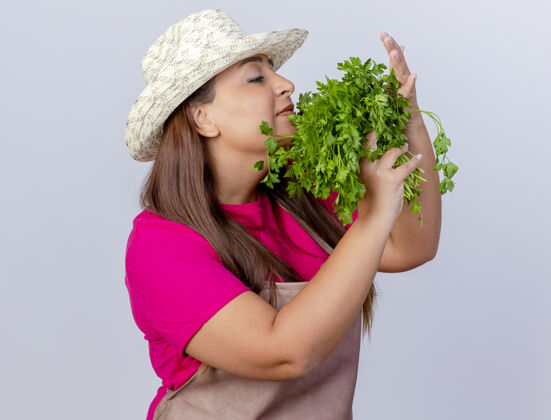 香气围裙戴帽子的中年园丁妇女站在白色的背景上 手里拿着新鲜的香草 散发着芳香中期草药女人