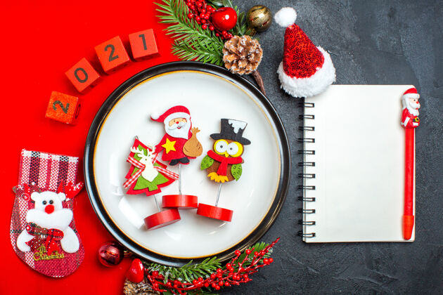 冷杉新年背景的俯视图 带有餐盘装饰配件杉木树枝和数字圣诞袜放在一个红色餐巾笔记本上 黑色桌子上有钢笔树枝盘子钢笔