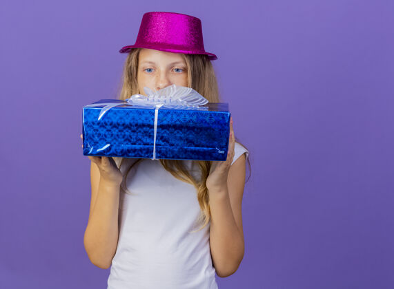 看漂亮的小女孩戴着节日礼帽拿着礼盒看着它兴致勃勃 生日派对的概念站在紫色的背景上盒子举行生日