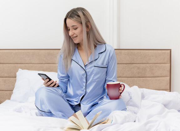 杯子穿着蓝色睡衣的年轻美女坐在床上 用智能手机喝着咖啡 在卧室的室内灯光背景下微笑着女士年轻咖啡