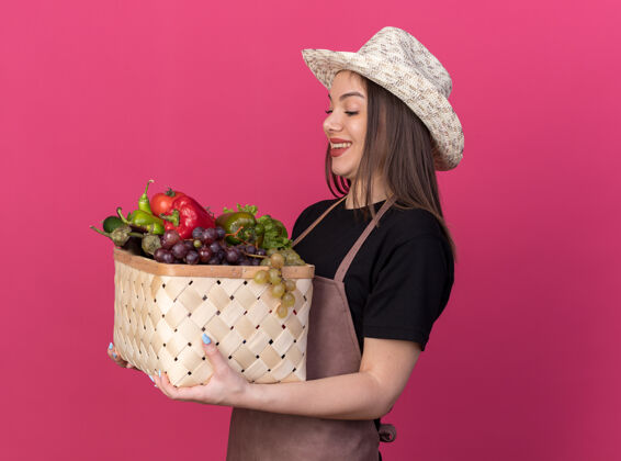 女微笑着美丽的白人女园丁戴着园艺帽拿着和看着蔬菜篮子隔离在粉红色墙上复制空间蔬菜花园帽子