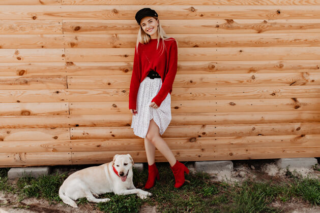 季节美丽的金发女郎穿着白色连衣裙和红色套头衫在木墙上摆姿势可爱的女孩和她的狗在户外感觉良好和快乐羊绒微笑肖像
