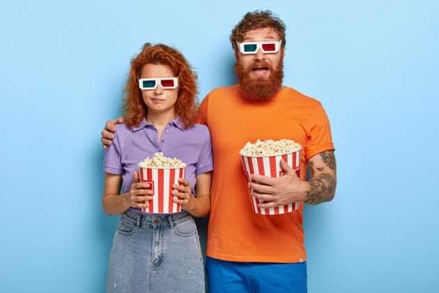 女朋友年轻的红发夫妇在3d影院里度过闲暇时光 吃爆米花 戴上特殊的眼镜以获得视觉效果 留着胡子的兴奋的家伙拥抱着表情无聊的可爱女友 不喜欢电影丈夫红发拥抱