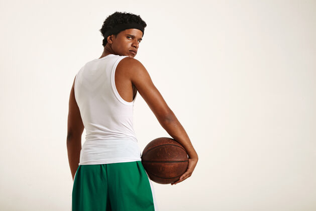 比赛严肃果断的年轻非洲裔美国篮球运动员 身穿白色和绿色制服 一个非洲裔短发 臀部举着一个棕色的旧篮球 回头看运动员年轻运动