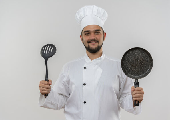 拿着身着厨师制服 面带微笑的年轻男厨师手拿着开槽汤匙和煎锅隔离在空白处煎炸制服开槽