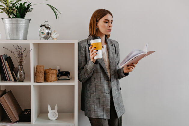 办公室年轻的女商人手里拿着一杯咖啡 痴迷于阅读 靠在架子上 架子上放着工作用具财务夹克行政人员