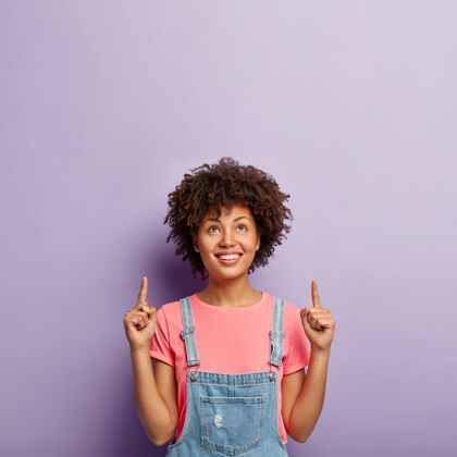 手指广告和促销概念可爱的卷发女人专注于上面 将食指指向复制空间 向上显示方向 穿着时尚的服装 隔离在紫色的墙上非洲微笑人