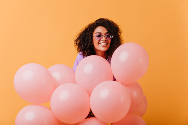 卷发戴着圆眼镜的年轻女子高兴地摆着粉红色的气球快乐的非洲生日女孩被隔离在黄色乐趣积极成人