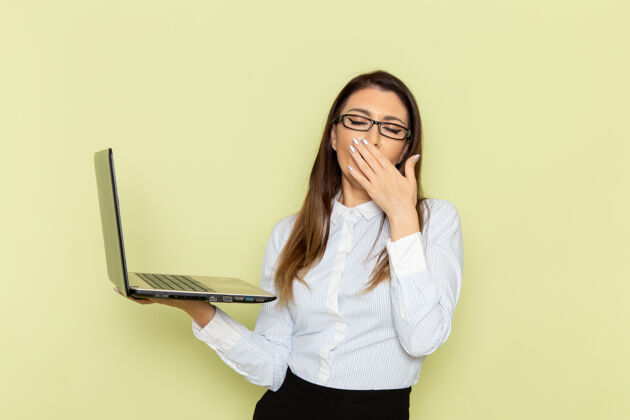 抱着穿着白衬衫黑裙子的女上班族手持笔记本电脑 打着哈欠站在浅绿色的墙上笔记本电脑工作商务